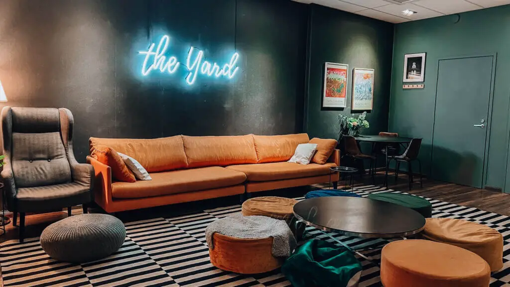 The Yard Hostel Helsinki Common Area features a long orange sofa, a chair next to it, a desk with three chairs and a mini table with pouffes around. There is a neon the yard sign on the wall above the sofa.