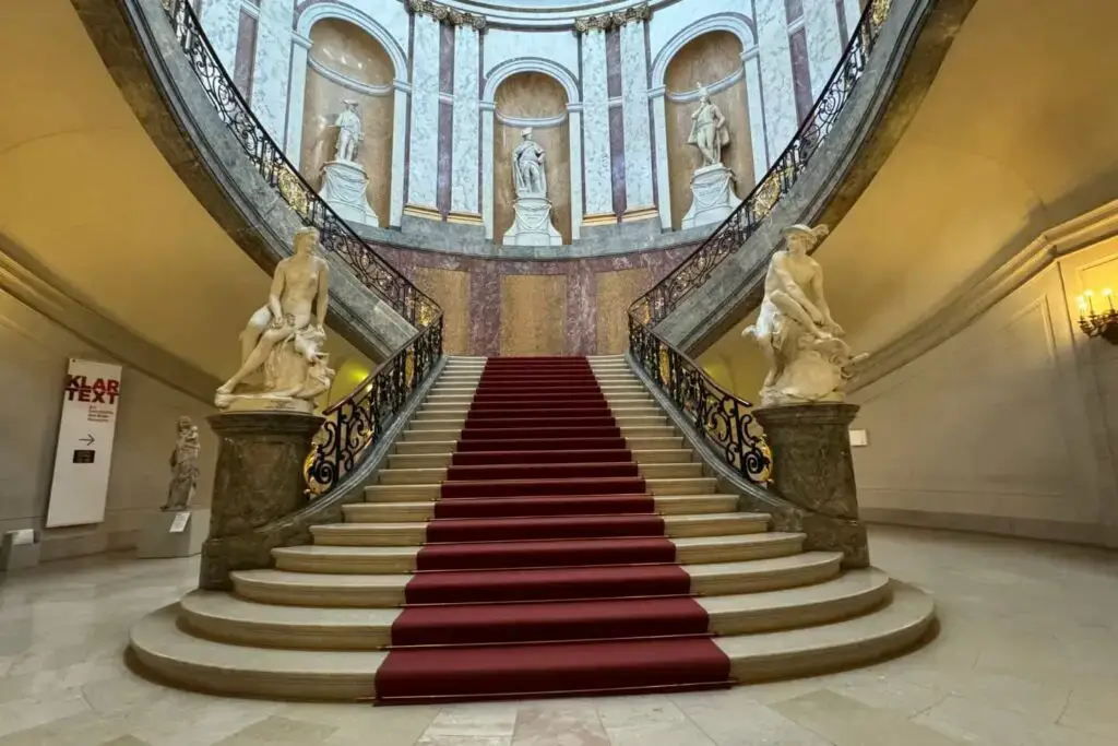 Bode Museum staircase part of the Free Museums Berlin Sunday. There is a grand staircase with a red carpet that goes to the right or the left with marble statues.