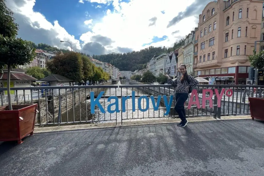 Is Karlovy Vary Worth Visiting? The karlovy vary sign along a bridge. there are picturesque hoses either side and Holly is posing in the middle.