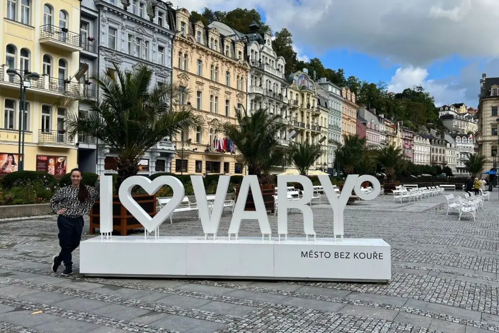 Is Karlovy Vary Worth Visiting? the karlovy vary sign with picturesqu houses behind it. Holly is posing on the left of the sign